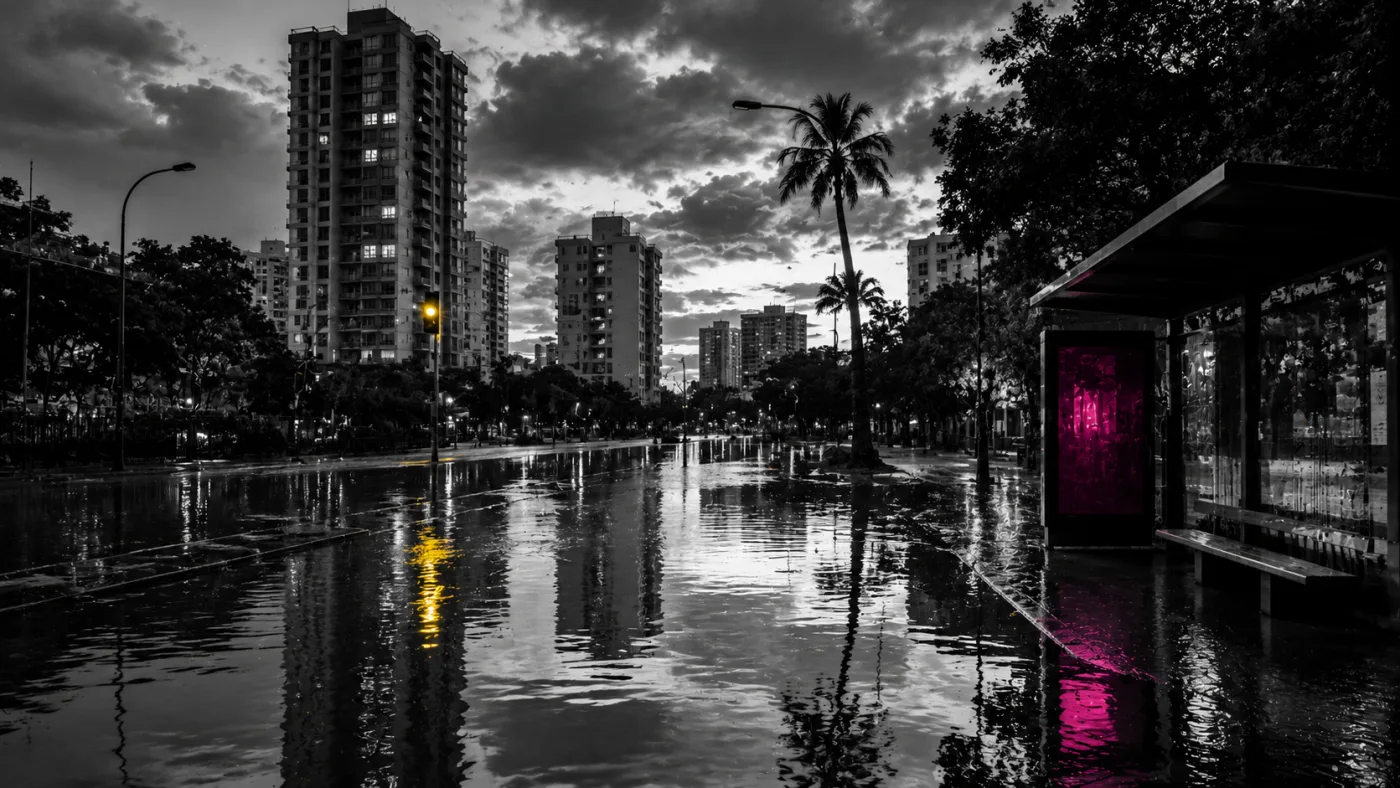 Rua alagada em cidade brasileira após chuva forte, com prédios e palmeiras ao fundo